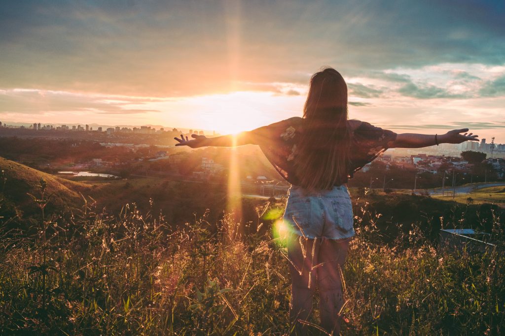 women with arms spread out looking over city with sun rising in background.